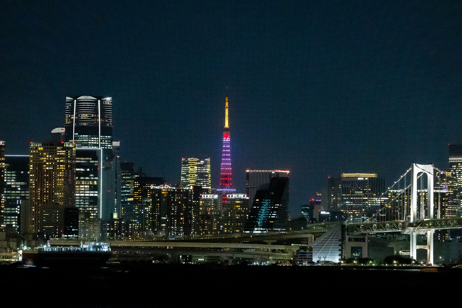 Tokyo Tower yang diterangi cahaya warna-warni. Di bagian depannya, Jembatan Rainbow Bridge.